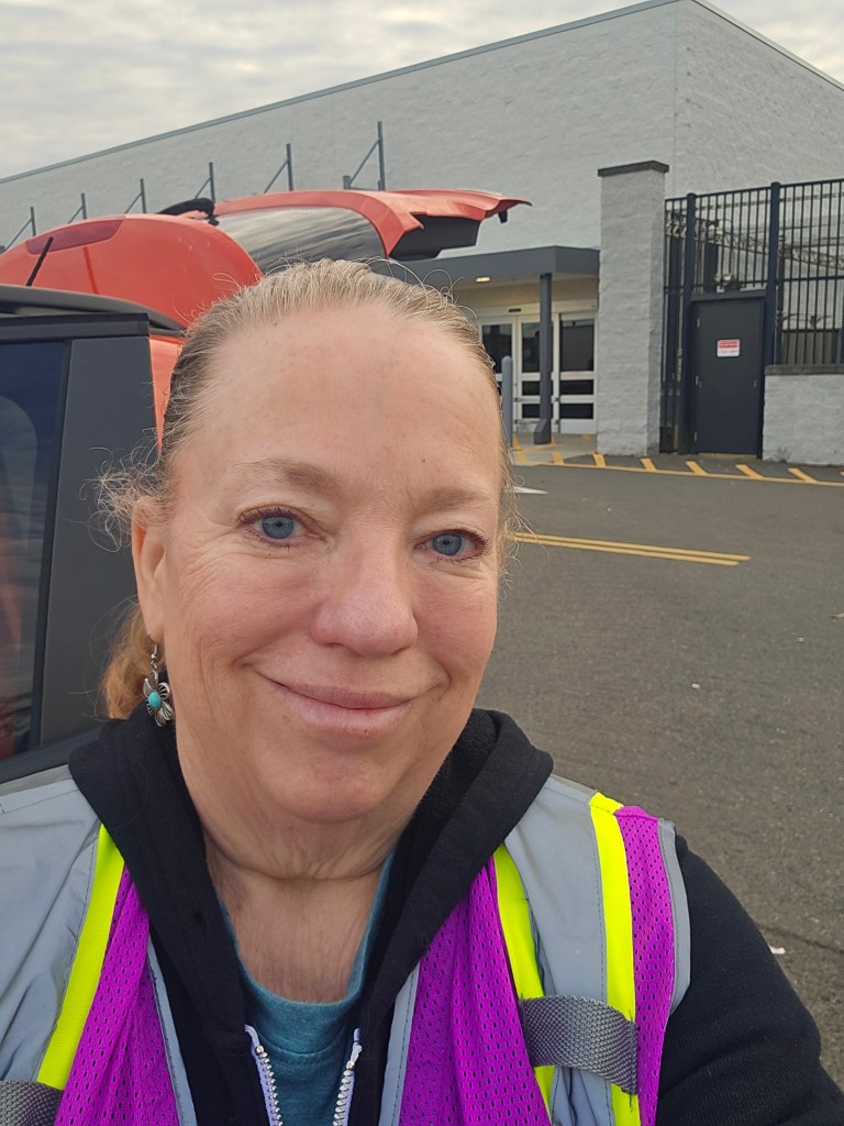 Smiling selfie of author Joy in a purple and yellow high-visibility safety vest, standing in a Walmart parking lot with her car’s hatchback open, ready for a Spark delivery.