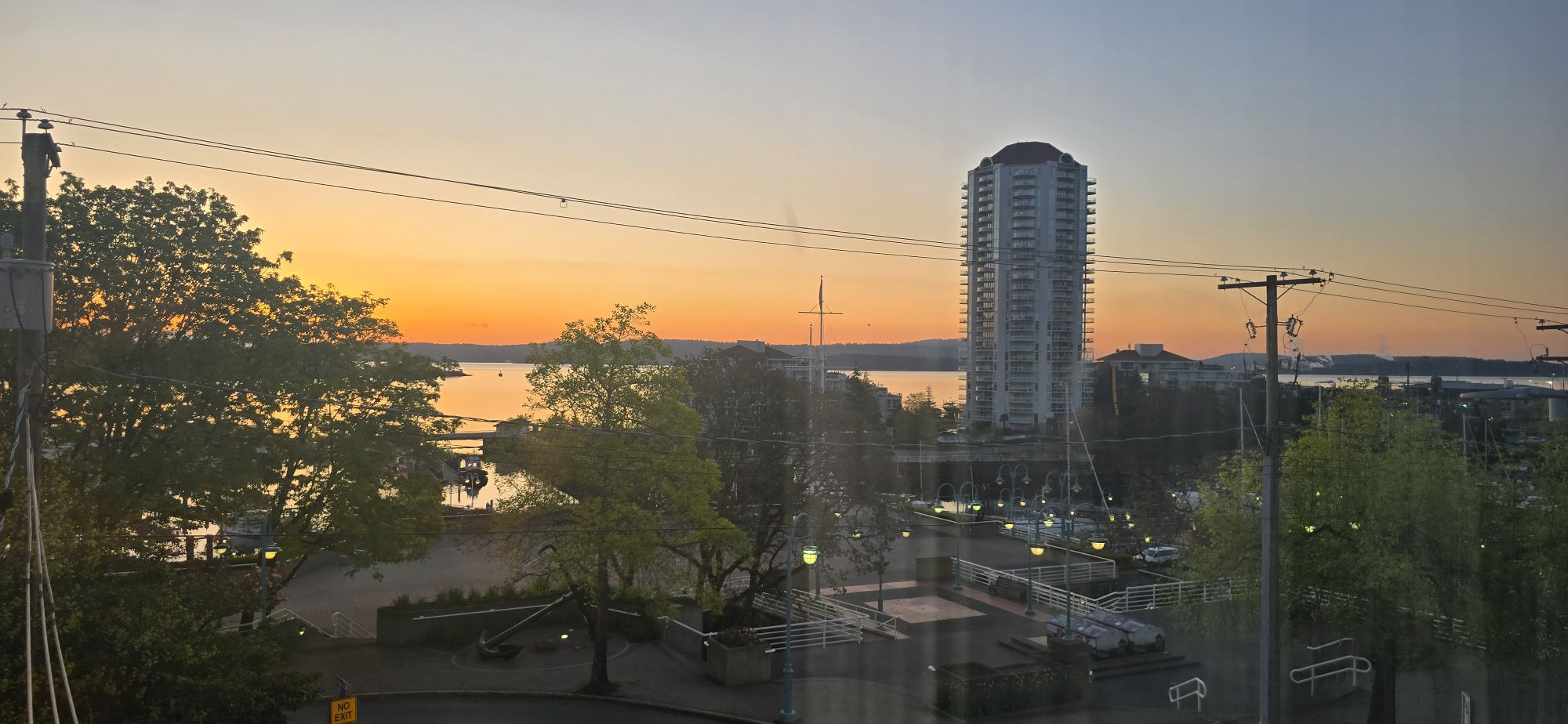 A high-angle view of the Nanaimo, Canada, harbor at sunrise. The eastern horizon is painted a dramatic gradient of deep orange, yellow, and blue. The calm water of the bay reflects the light, with small boats docked nearby. In the center-right is the prominent white high-rise Cameron Island tower. The view is framed by dense, bright green spring foliage and foreground utility wires, indicating a view from a window or hotel room above the streets.