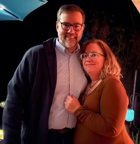 A candid nighttime photograph of Joy and Scott Beatty, smiling and embracing at a casual backyard wedding celebration. Joy wears a rust-colored long-sleeve shirt and glasses; Scott wears a patterned button-down, a dark cardigan, and glasses. Joy’s hands rest affectionately on Scott’s chest as they lean in together.