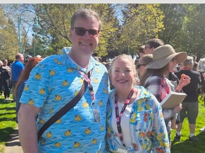 A bright, outdoor photo of Scott and Joy Beatty smiling together at a sunny public event in Nanaimo. Scott is wearing a blue short-sleeved shirt with a yellow rubber duck pattern and sunglasses. Joy is wearing a white top with a colorful floral-patterned overshirt and a red-and-white Canada-themed lanyard. A crowd of people and green trees are visible in the background.