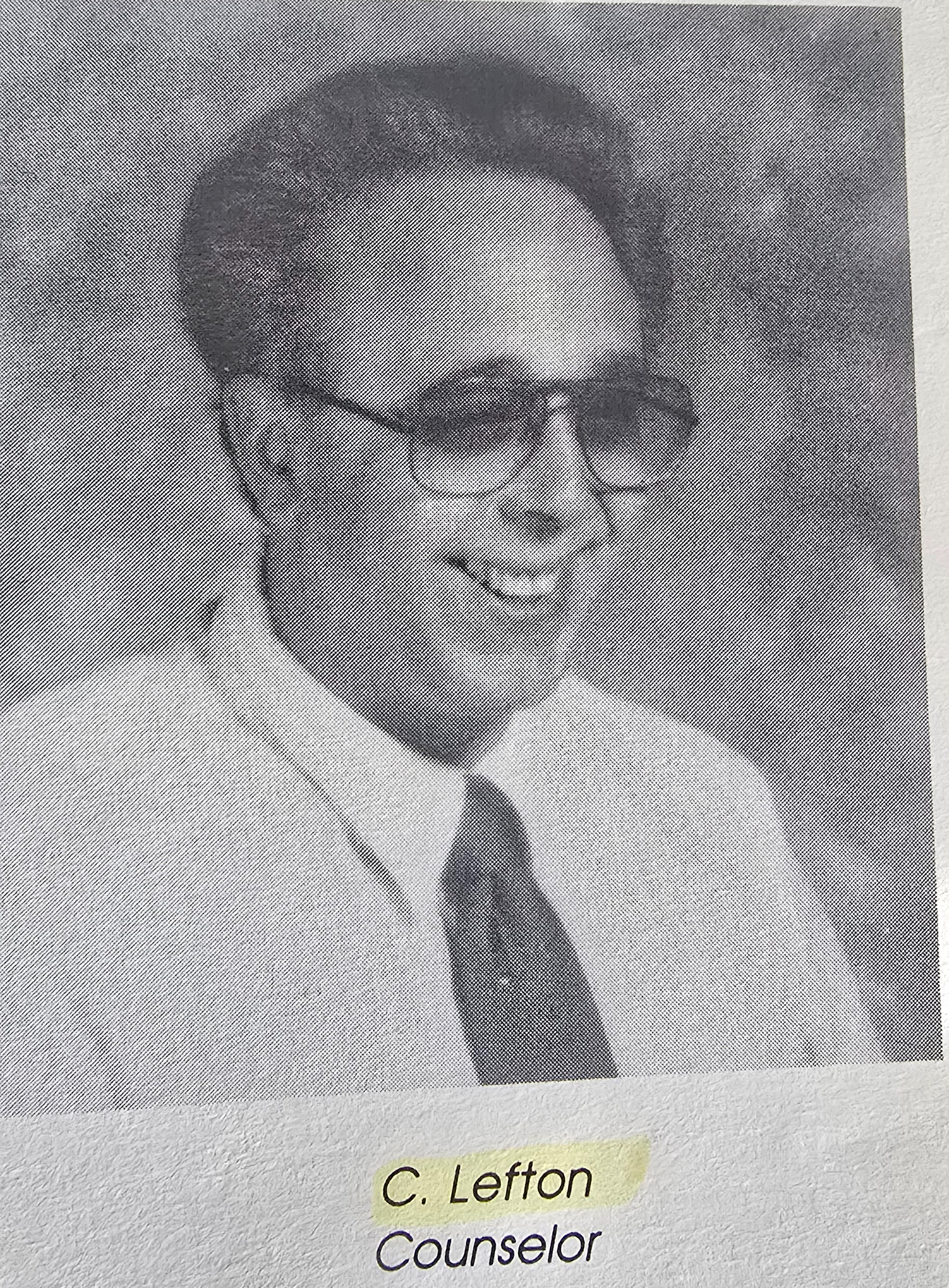 A high-contrast, black-and-white headshot of a middle-aged man with dark hair, balding at the crown, wearing thin-rimmed eyeglasses, a light-colored button-down shirt, and a dark tie. He is looking off to the right of the frame with a wide, toothy smile. Below the photograph, text printed on the yearbook page reads, C. Lefton, Counselor
