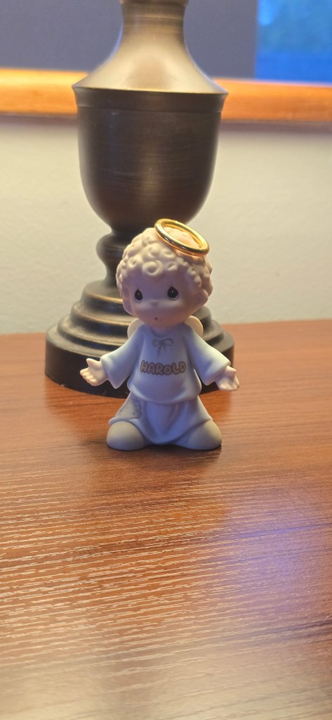 A vintage Precious Moments porcelain figurine of a small angel boy with curly hair and a gold halo. He stands with his arms outstretched and palms upturned. On the chest of his light blue robe, the name "HAROLD" is printed in soft brown decorative script. He is sitting on a dark wood surface in front of a lamp.