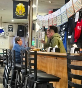 An elderly woman with short dark hair and a blue sweater sits on a tall black barstool at a wood-paneled bar. She is handing cash to a male bartender in a green hoodie. In the background, there are pool cues, a "Hefeweizen" banner, and several rows of colorful pull-tab games hanging above the bar.