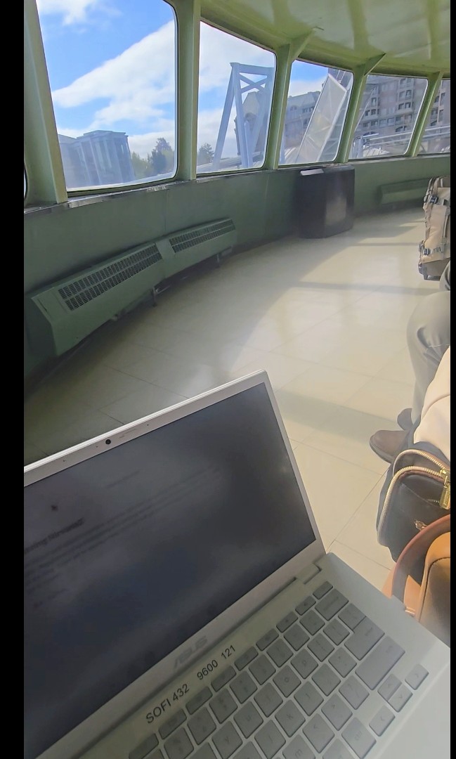 A first-person perspective from the front row of the Coho ferry. Joy’s knees serve as a makeshift desk for her trusty white ASUS Chromebook, which is open and active as she writes. Through the large, panoramic ferry windows, the bright morning sky and the shoreline are visible. The sun streams into the cabin, illuminating the pale green interior and the "laptop-on-knees" reality of a writer in motion.